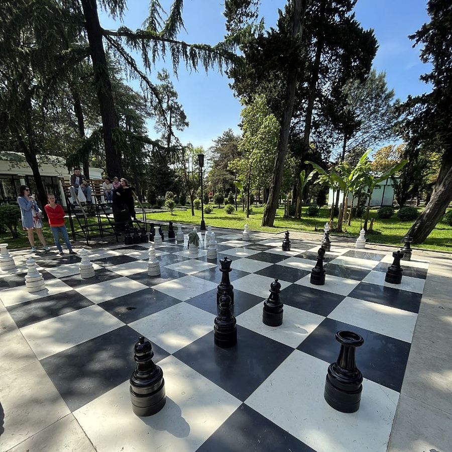 A giant outdoor chessboard with waist-high pieces in a tree-lined park.