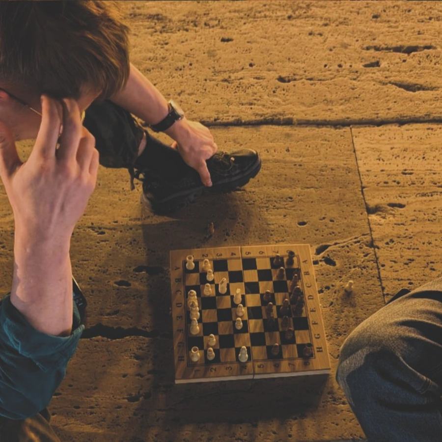 Igor Shatsev thinking over a travel chess set on a stone floor, hand to his head, his opponent at the edge of the frame.