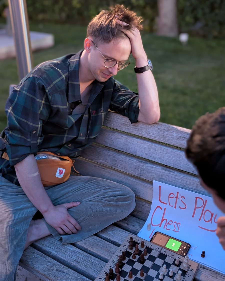 Igor Shatsev on a wooden bench at the Cascade in Yerevan, hand in his hair, smiling down at a chess board and a phone clock; a hand-lettered “Let’s Play Chess” sign beside him, his opponent at the edge of the frame.
