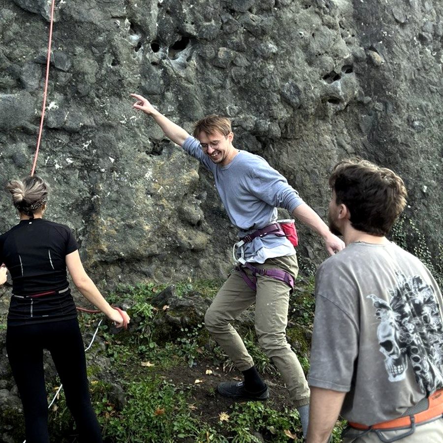 Igor Shatsev at the foot of an outdoor crag, laughing mid-gesture, two climbing partners nearby with their backs to the camera.
