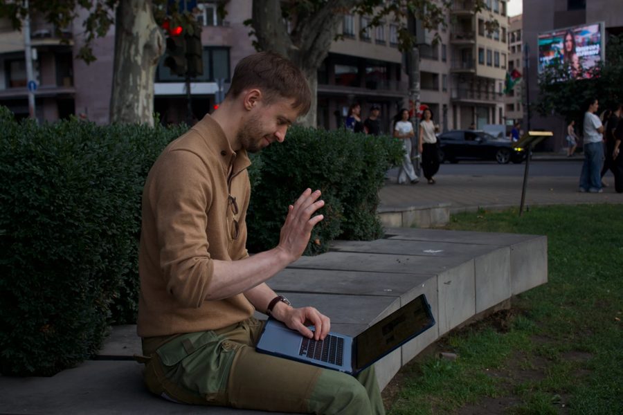 Igor Shatsev on a stone bench in a Yerevan square, laptop on his knees, waving at the screen at the start of an online lesson.