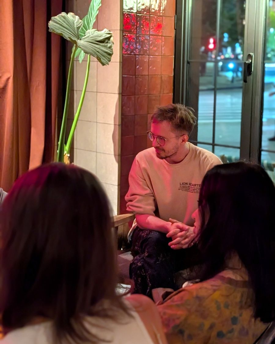 Igor Shatsev leading an English conversation club in a café at night, listening to a participant; two others sit with their backs to the camera.